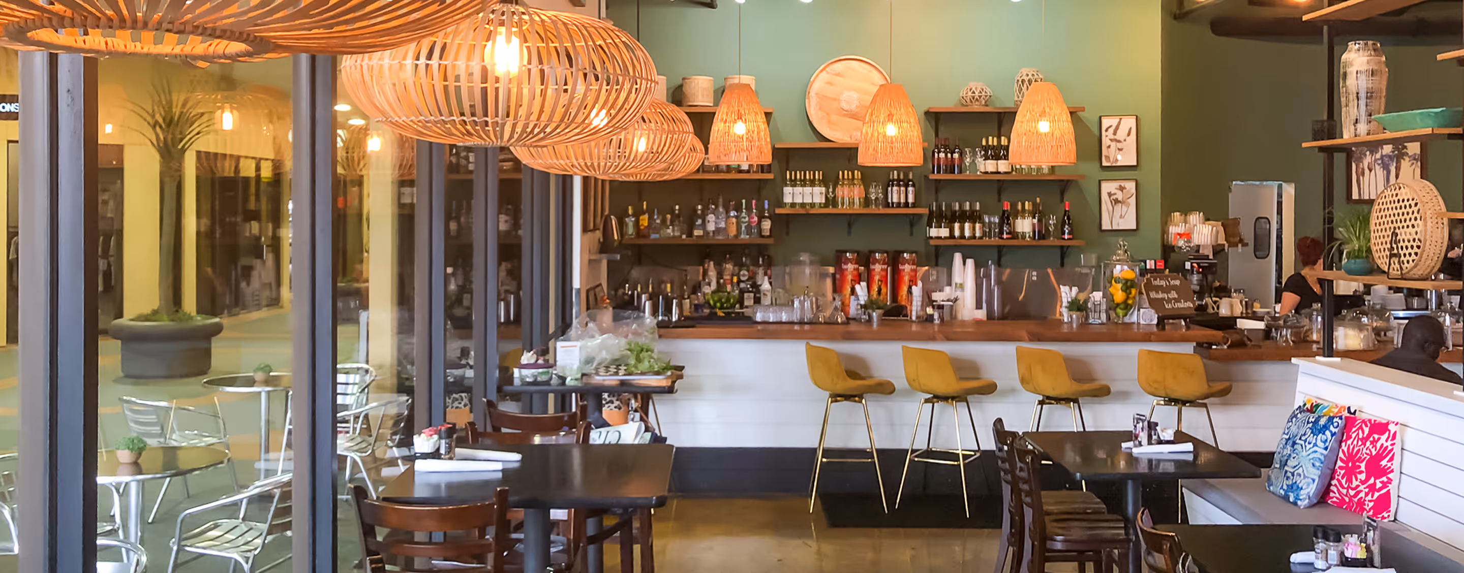 Interior of a cozy cafe with wooden tables, colorful cushions, yellow bar stools, and hanging wicker light fixtures.
