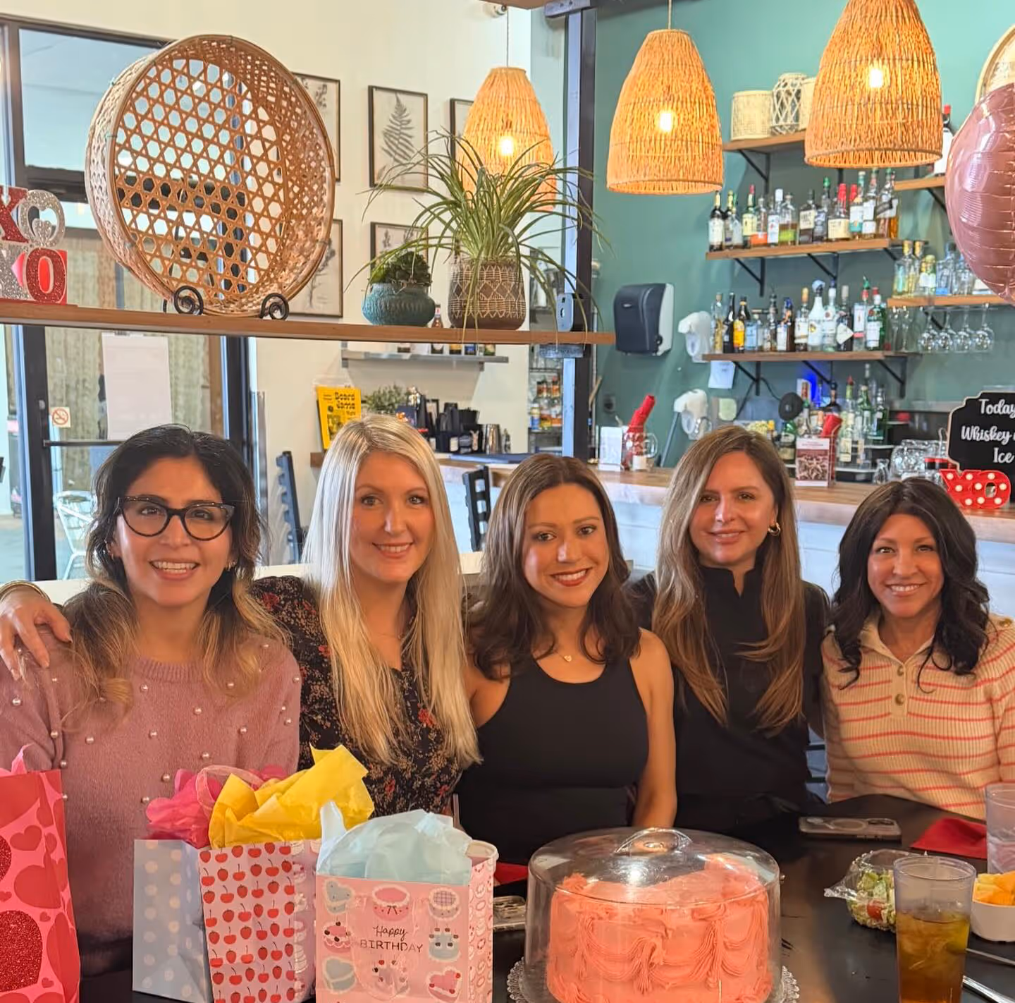 Five smiling women seated at a table with birthday gift bags and a pink frosted cake in a cozy decorated cafe.