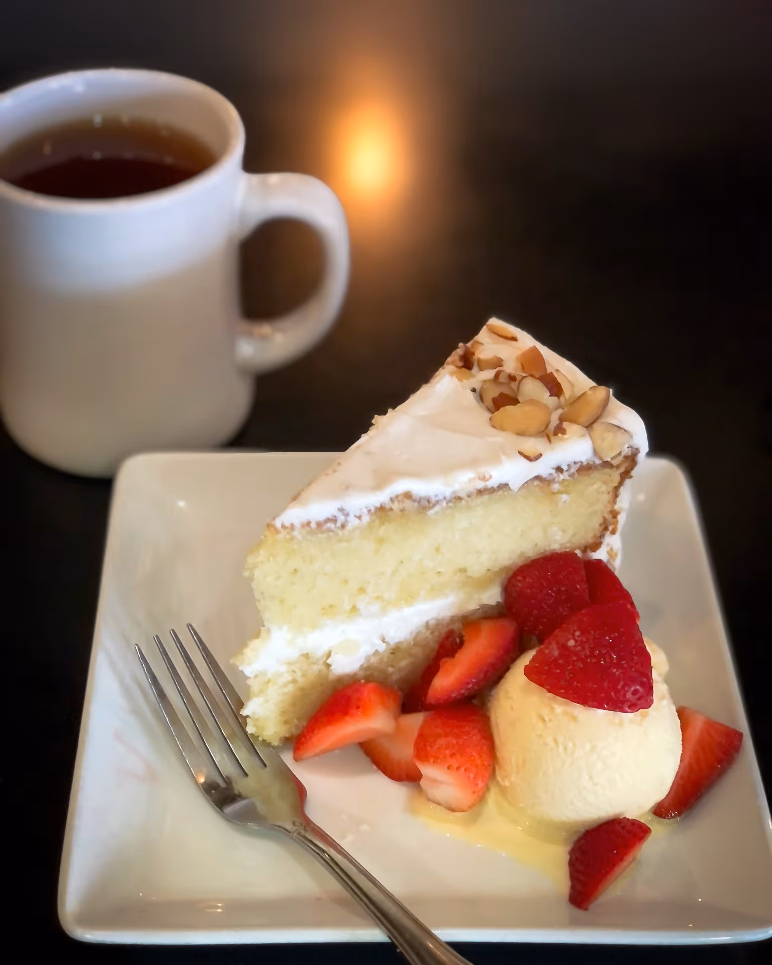 Slice of vanilla cake with white frosting and nuts, served with vanilla ice cream and sliced strawberries on a white square plate with a fork, and a cup of tea on a dark surface.