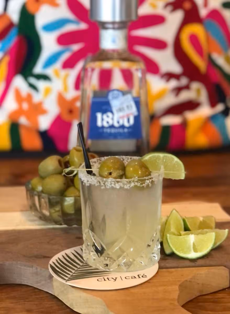 Cocktail glass with salted rim garnished with olives and a lime wedge, placed on a coaster labeled 'city | café' with a bottle of 1800 tequila and lime wedges in the background.