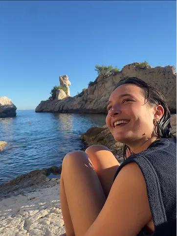 Jeune femme souriante assise sur une roche près de la mer avec des falaises rocheuses et un ciel bleu clair en arrière-plan.