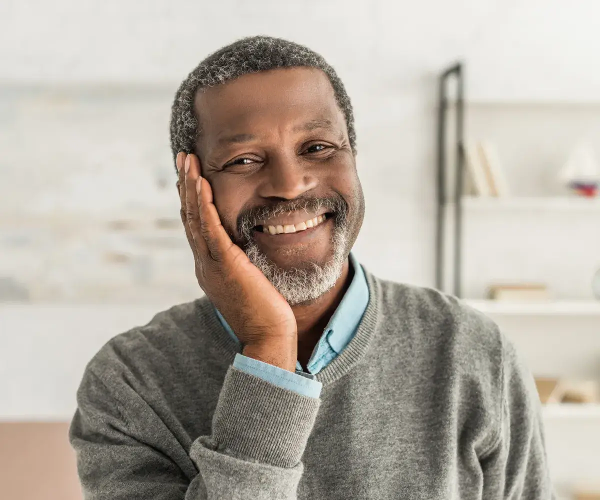 A smiling man in a gray sweater with his hand on his face.