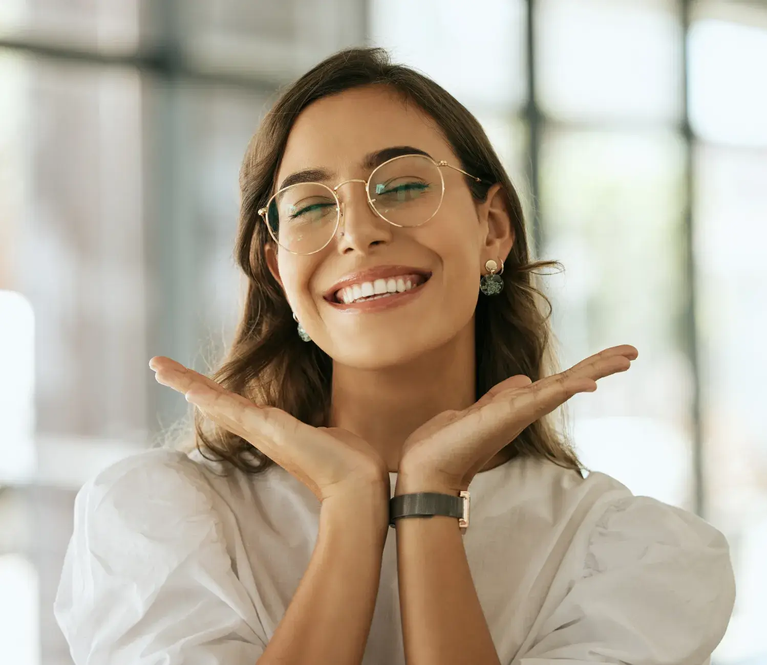 A woman wearing glasses is smiling and holding her hands together.