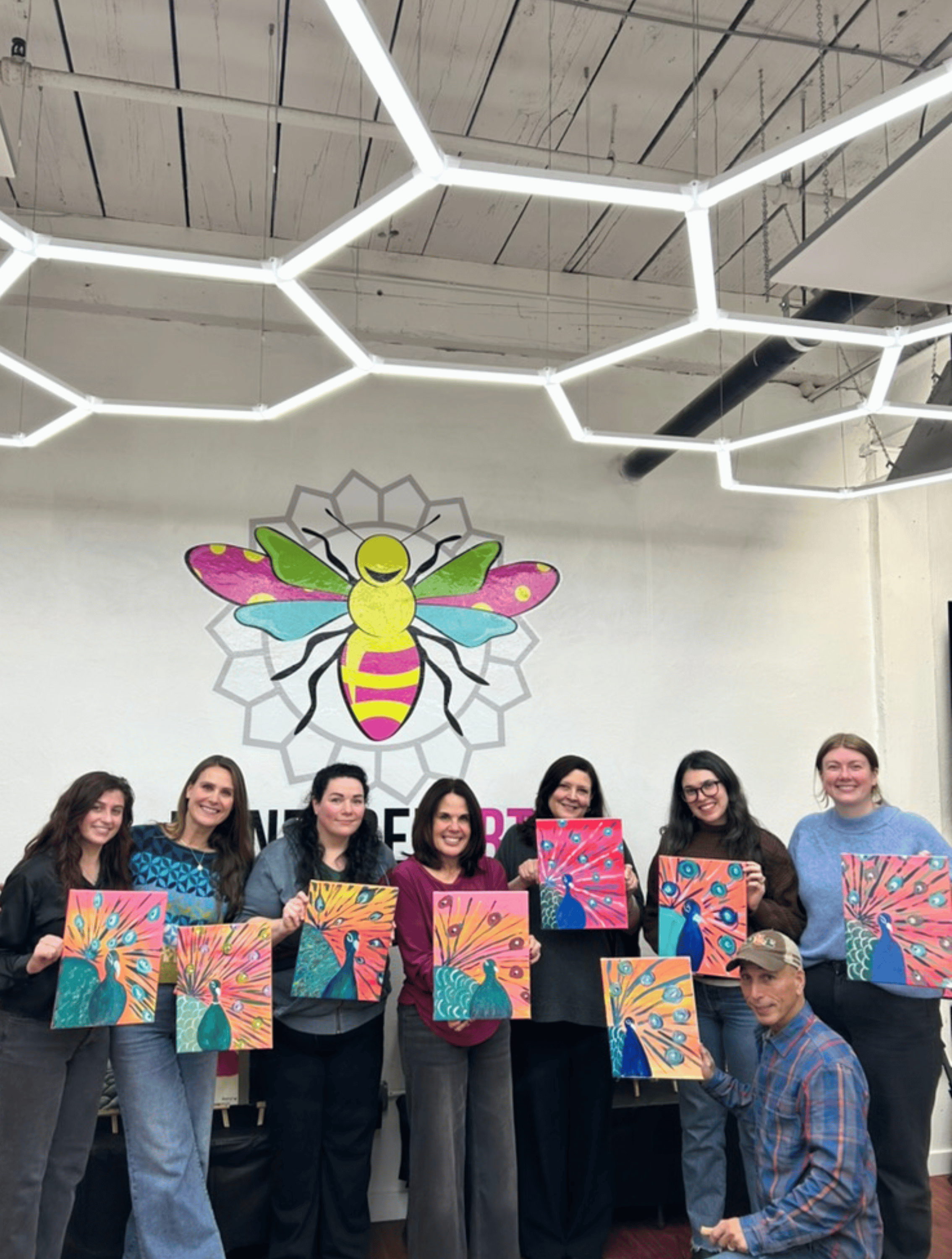 Group of sixteen smiling adults seated and standing indoors holding paintings of various cats and dogs.