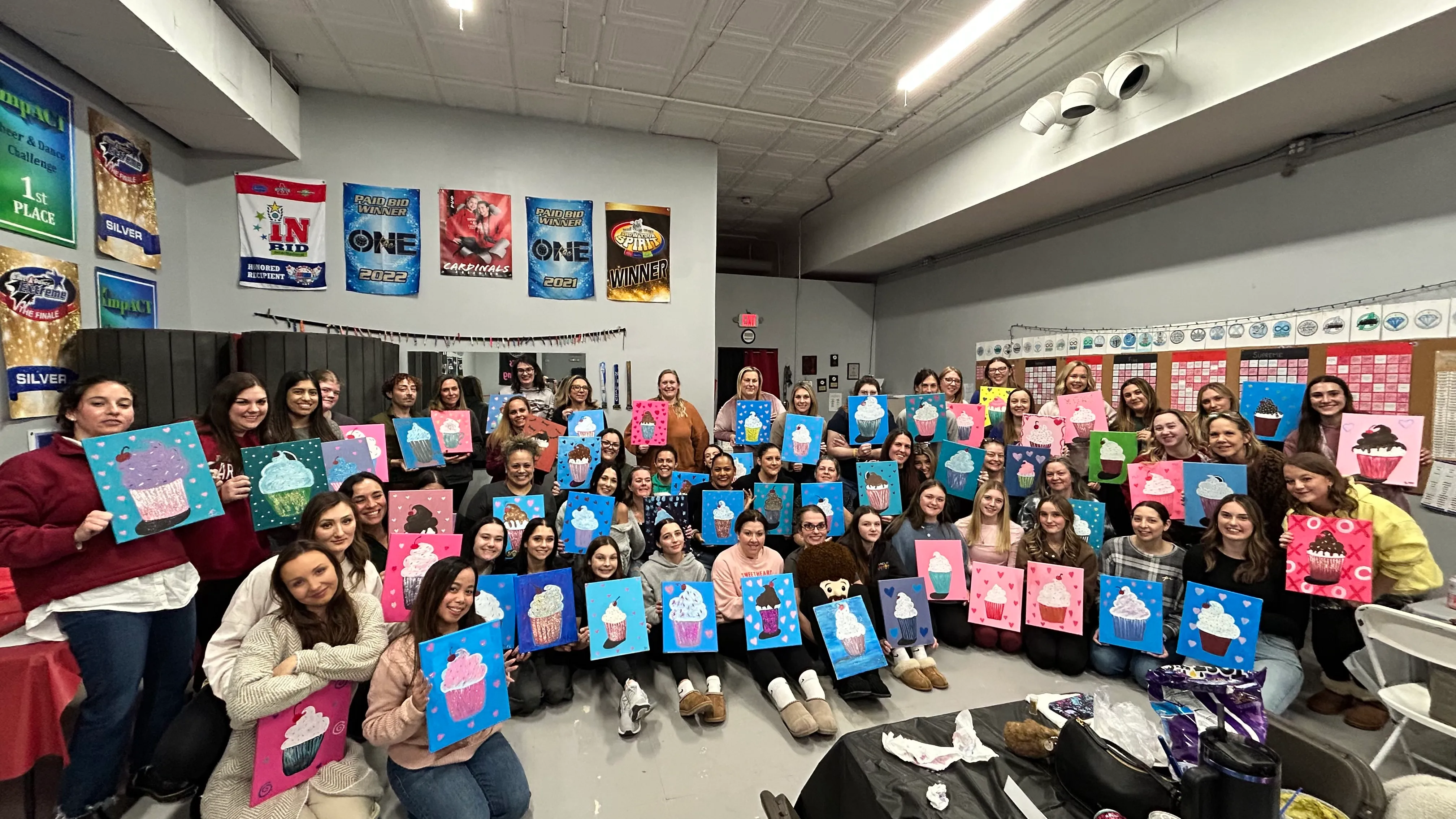 Large group of women holding cupcake paintings in a room with banners on the wall at a mobile paint party.