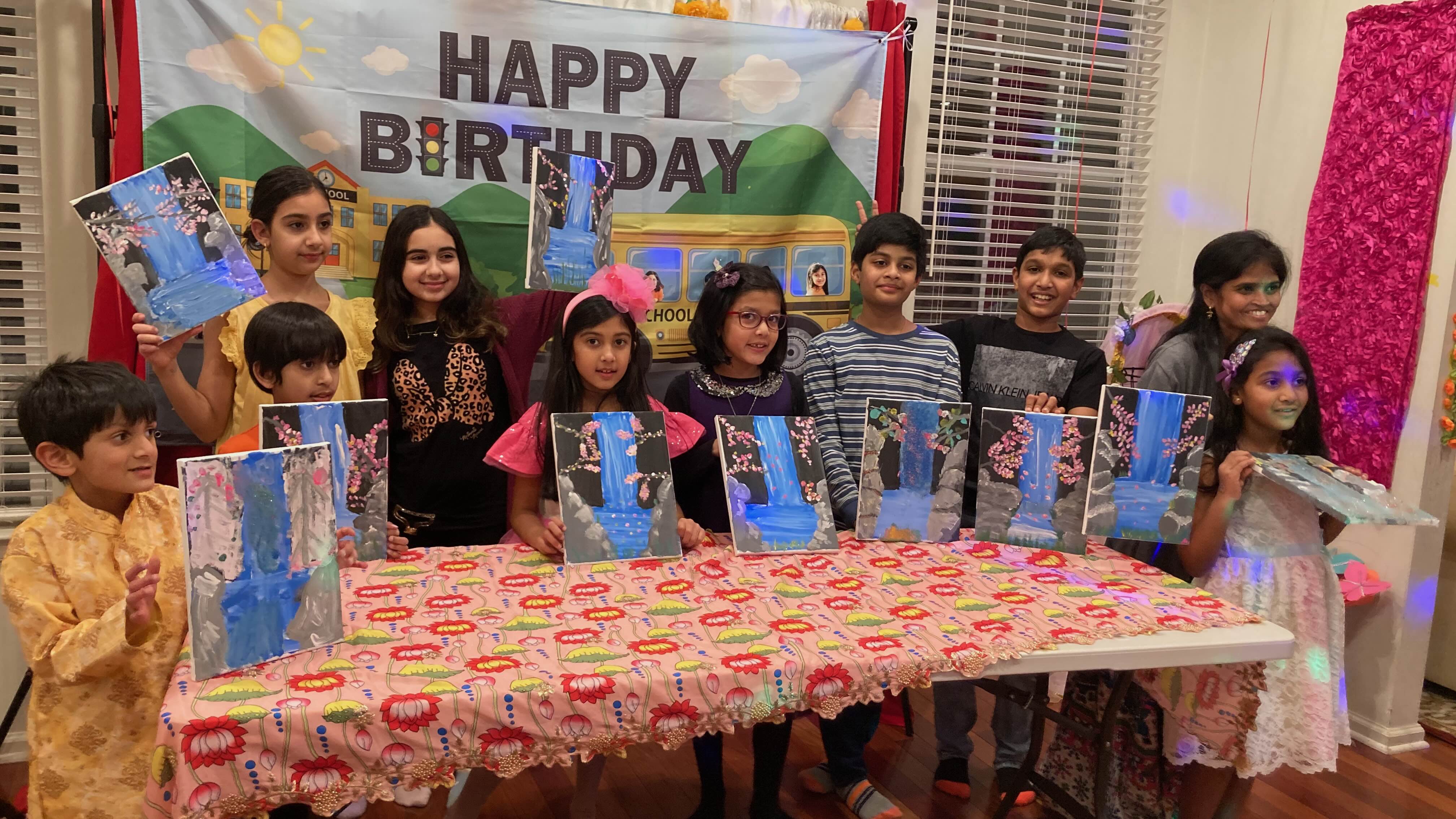 Group of children standing behind a table holding paintings of waterfalls with pink flowers, under a 'Happy Birthday' banner.