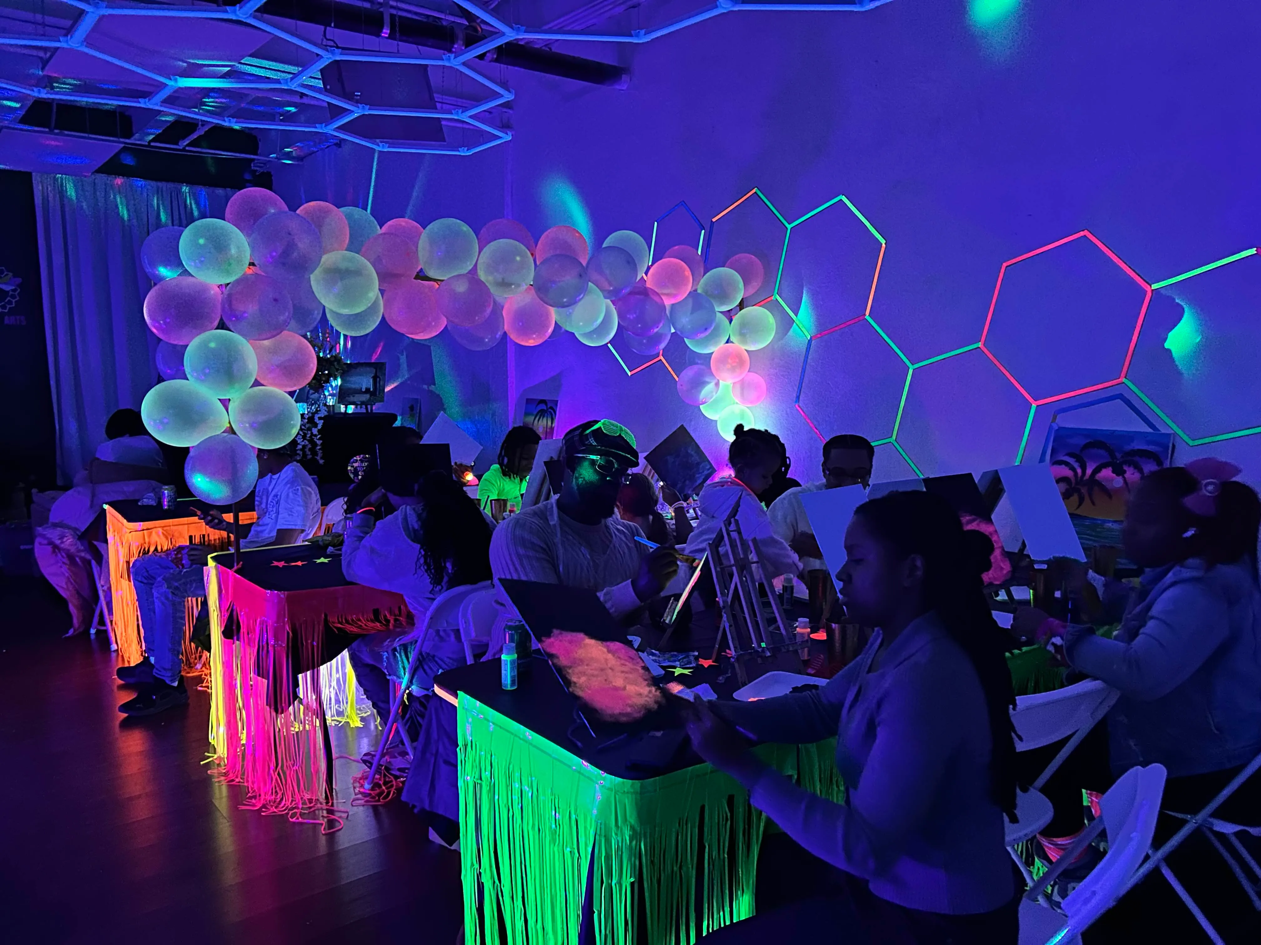 Group of people painting at tables decorated with glowing fringe and balloons under blacklight in an art studio.