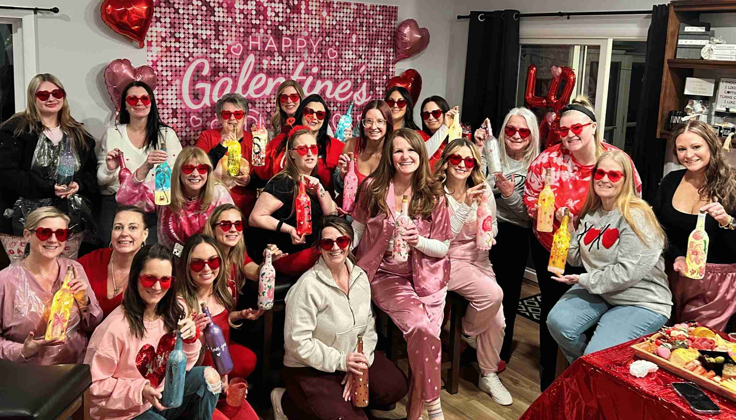 Group of women wearing heart-shaped sunglasses holding decorated bottles, posing in front of a pink 'Happy Galentine's' backdrop with heart-shaped balloons.