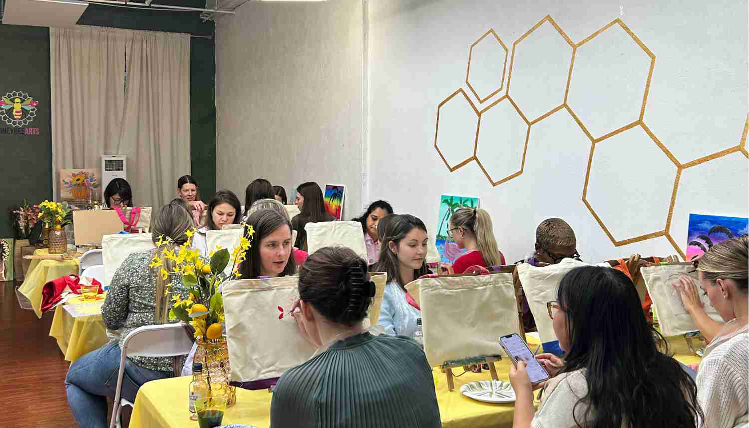 Group of diverse people posing indoors, each holding colorful paintings of sunrises reflecting on water, with a large bee mural on the wall behind them.