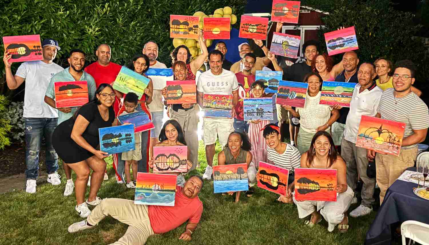 Group of people outdoors at night holding colorful paintings of guitars and sunsets, smiling and posing on grass.