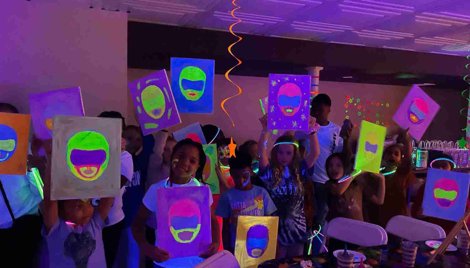 Group of people painting at tables decorated with glowing fringe and balloons under blacklight in an art studio.