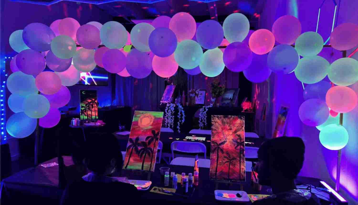 Dimly lit room with colorful balloons overhead, two people painting tropical sunset scenes with palm trees on canvases at a table.