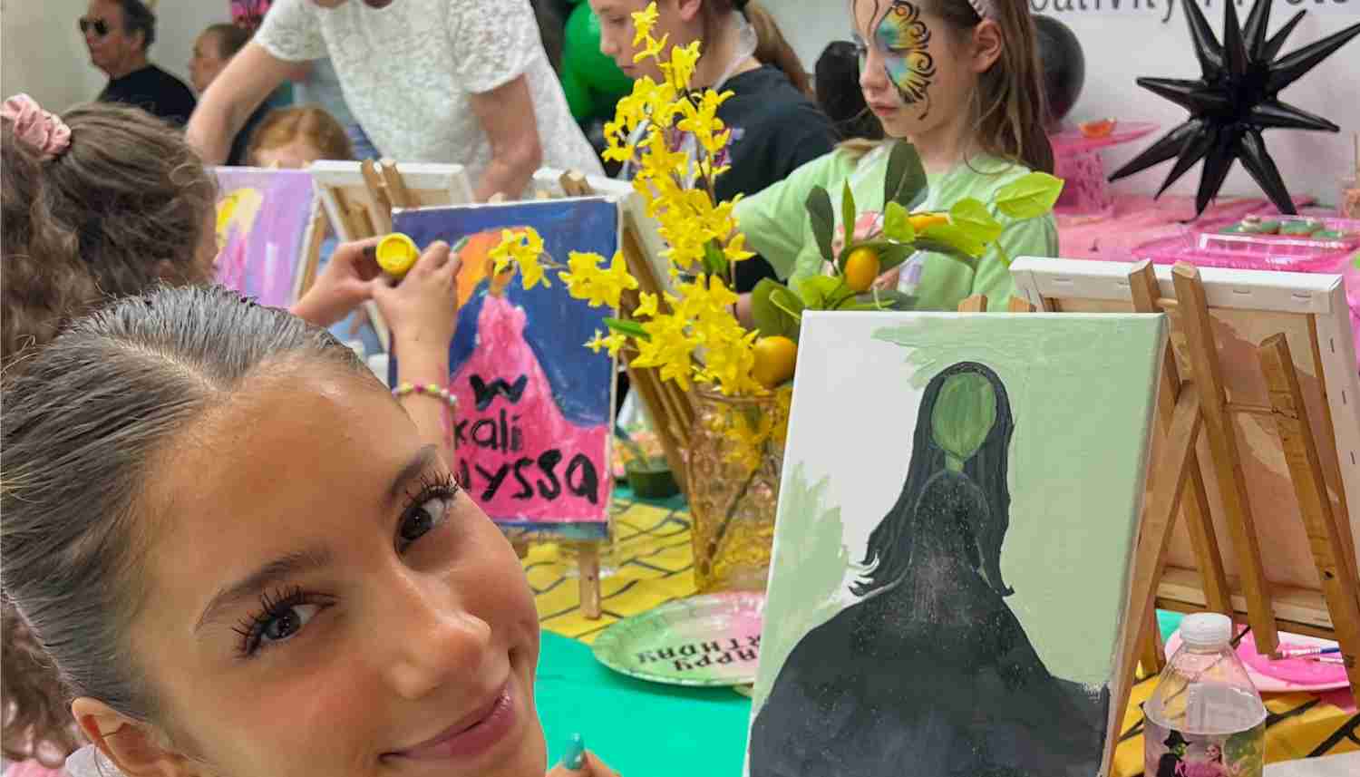 Smiling girl holding a painting of a woman in a black dress with a green face at a colorful art party with other children painting and face painted girl in the background.