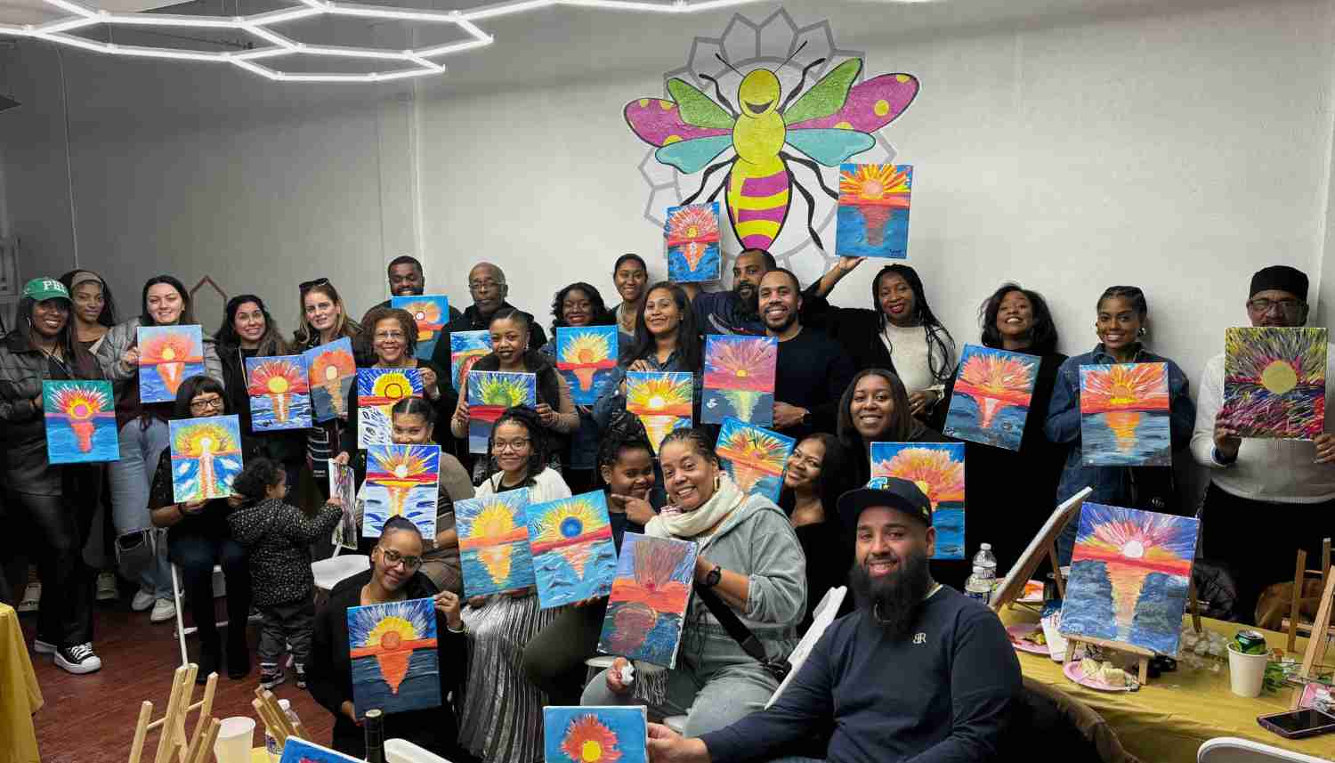 Group of diverse people posing indoors, each holding colorful paintings of sunrises reflecting on water, with a large bee mural on the wall behind them.