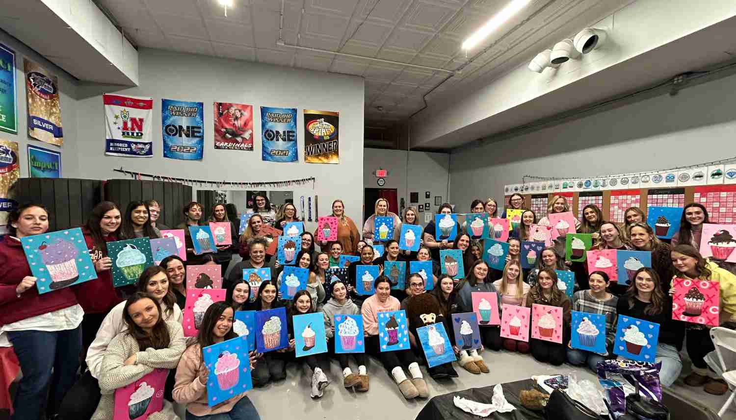Group of people posing indoors holding colorful paintings of cupcakes with various backgrounds.