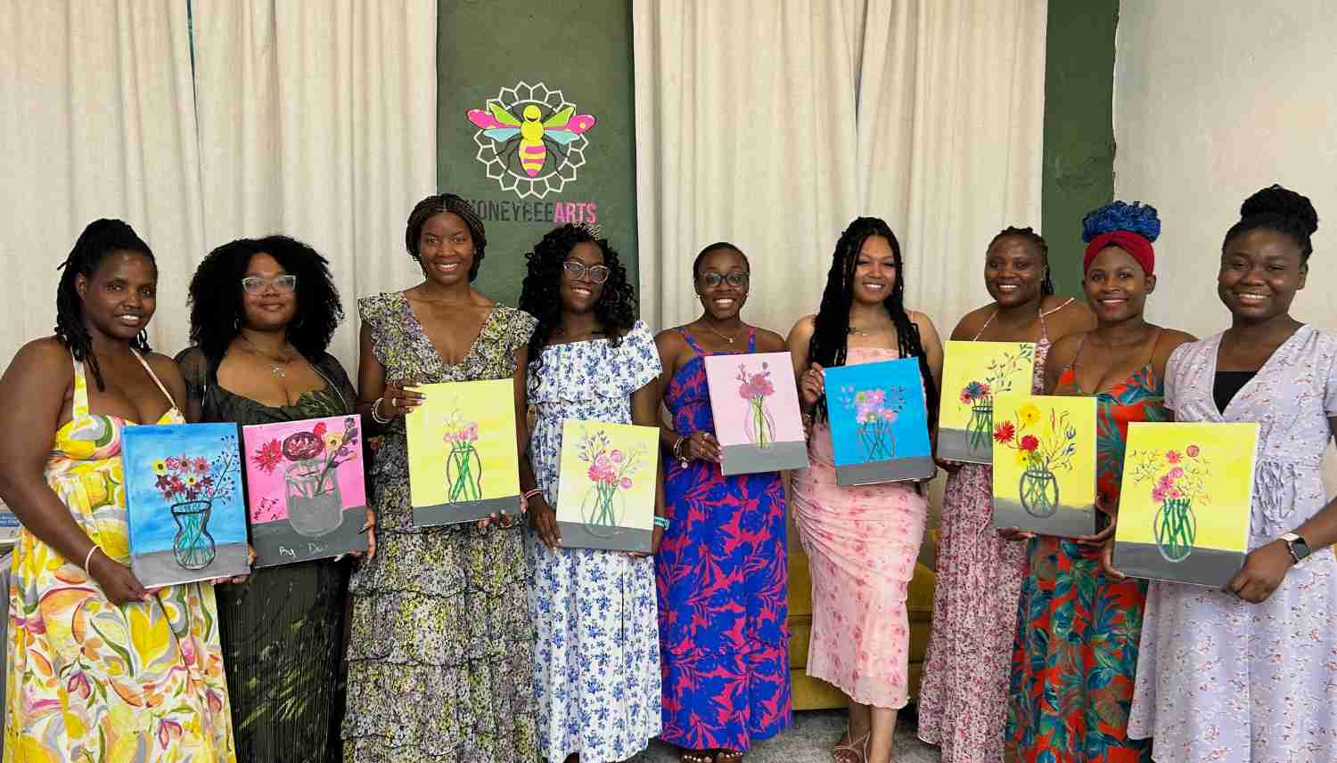 Nine women standing side by side holding paintings of flower bouquets in vases, smiling in a room with a green banner displaying a bee logo behind them.