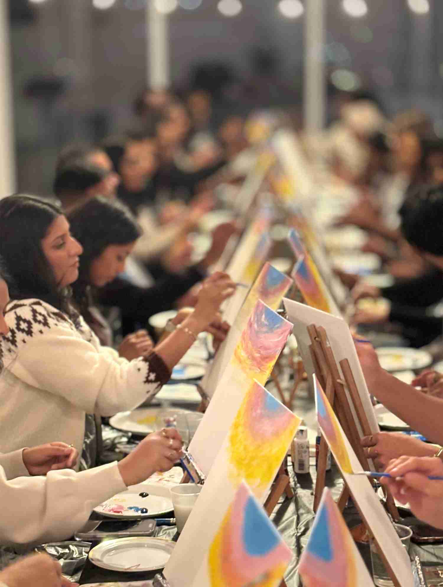 Group of people painting on canvases at easels in a room decorated with yellow flowers and art supplies.