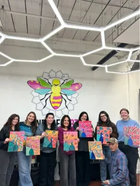 Group of sixteen smiling adults seated and standing indoors holding paintings of various cats and dogs.