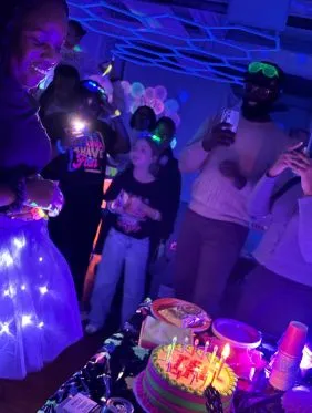Group of people celebrating a birthday indoors with colorful lights, featuring a cake with candles on a table.