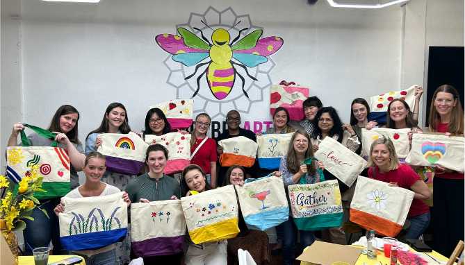 Large group of people posing indoors, each holding a painting of a colorful cupcake on a bright background.