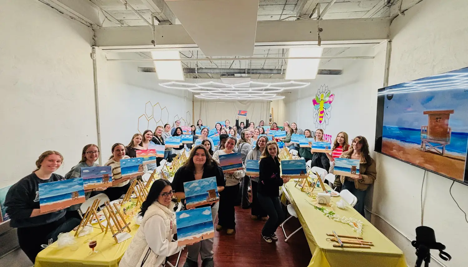 Group of people in a bright art studio holding up paintings of a beach scene with ocean and lifeguard tower.