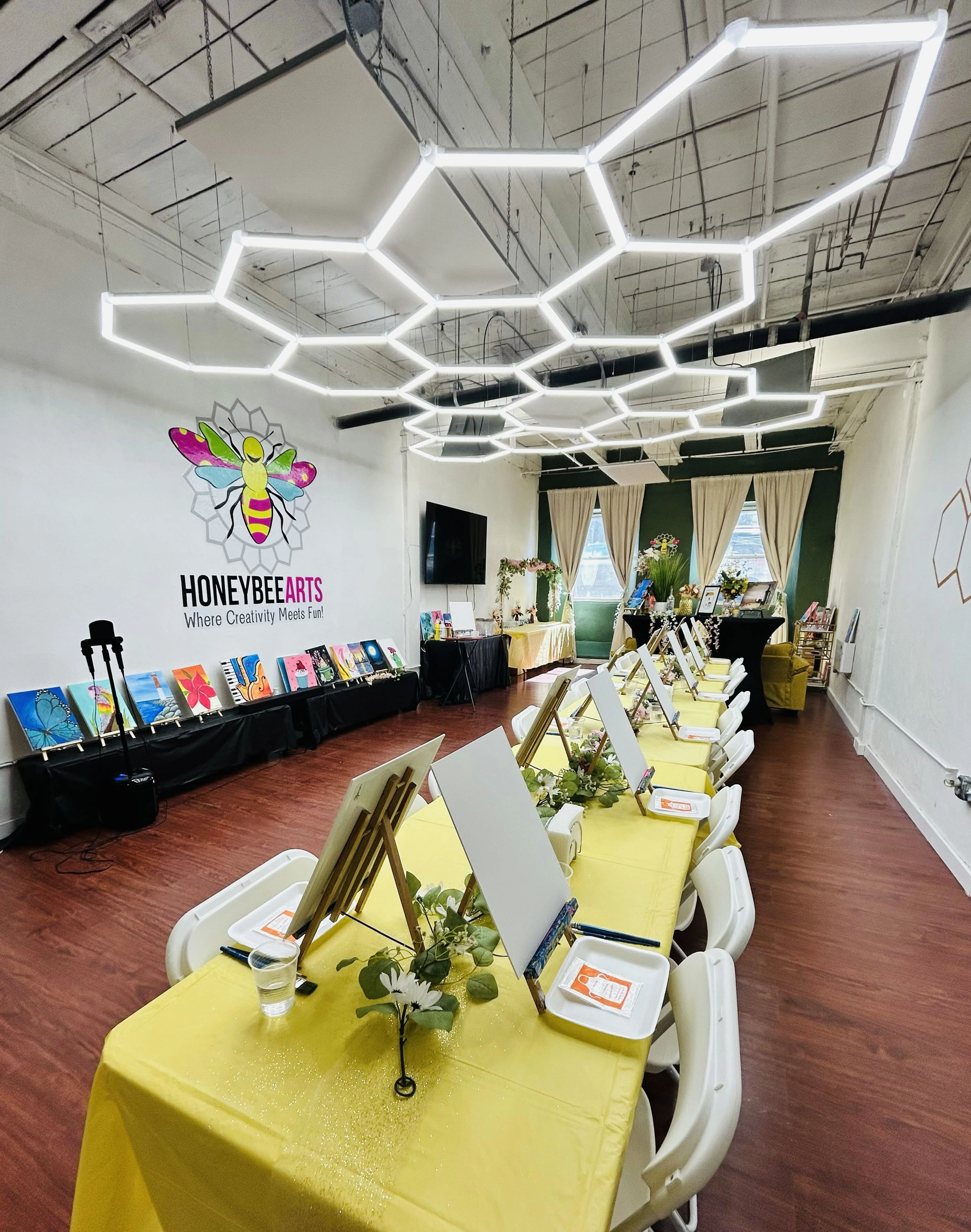 Art studio with long tables covered in yellow cloth, white chairs, easels with blank canvases, and decorative flowers under geometric LED ceiling lights.