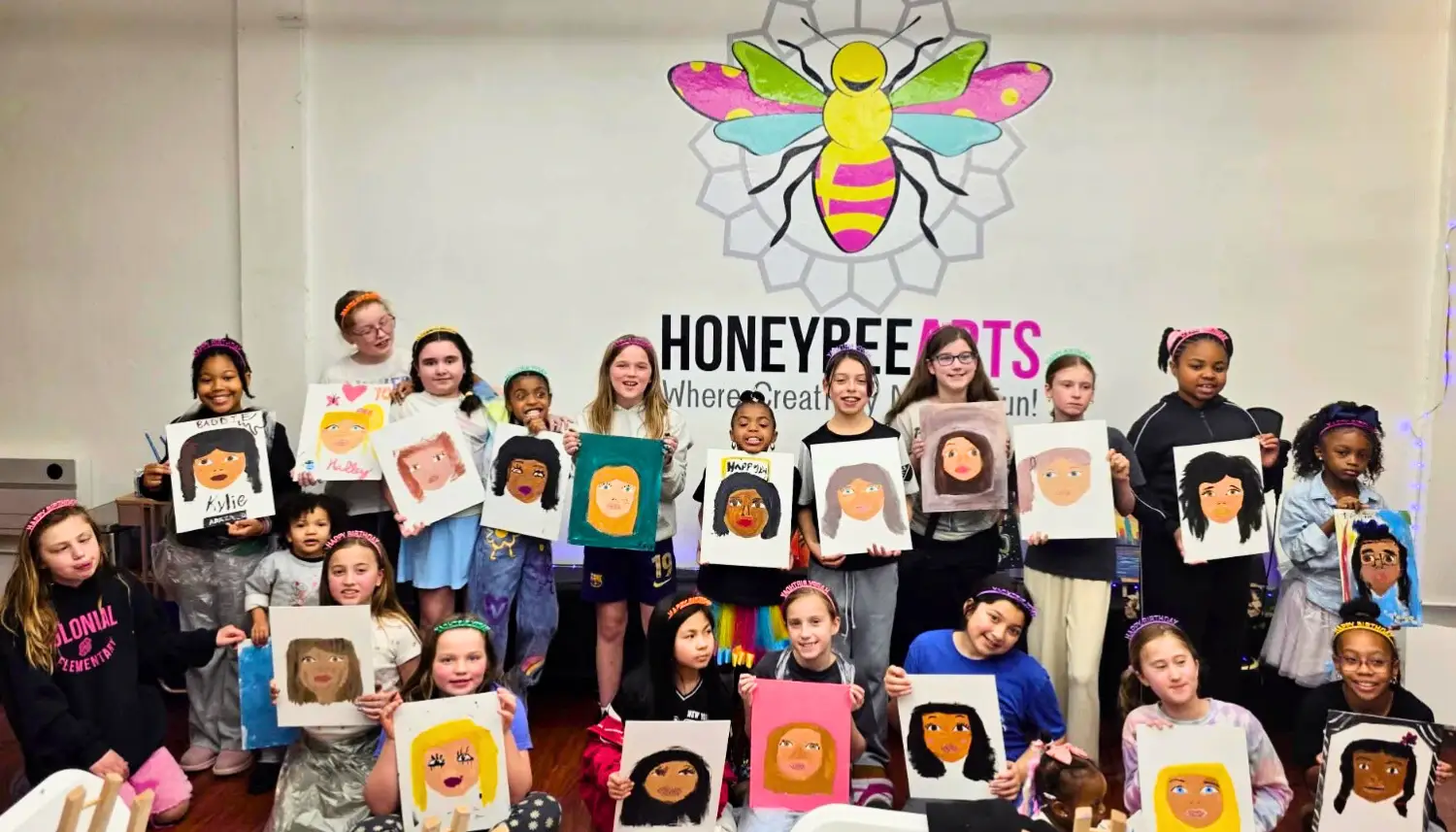 Group of children standing behind a table holding paintings of waterfalls with pink flowers, under a 'Happy Birthday' banner.