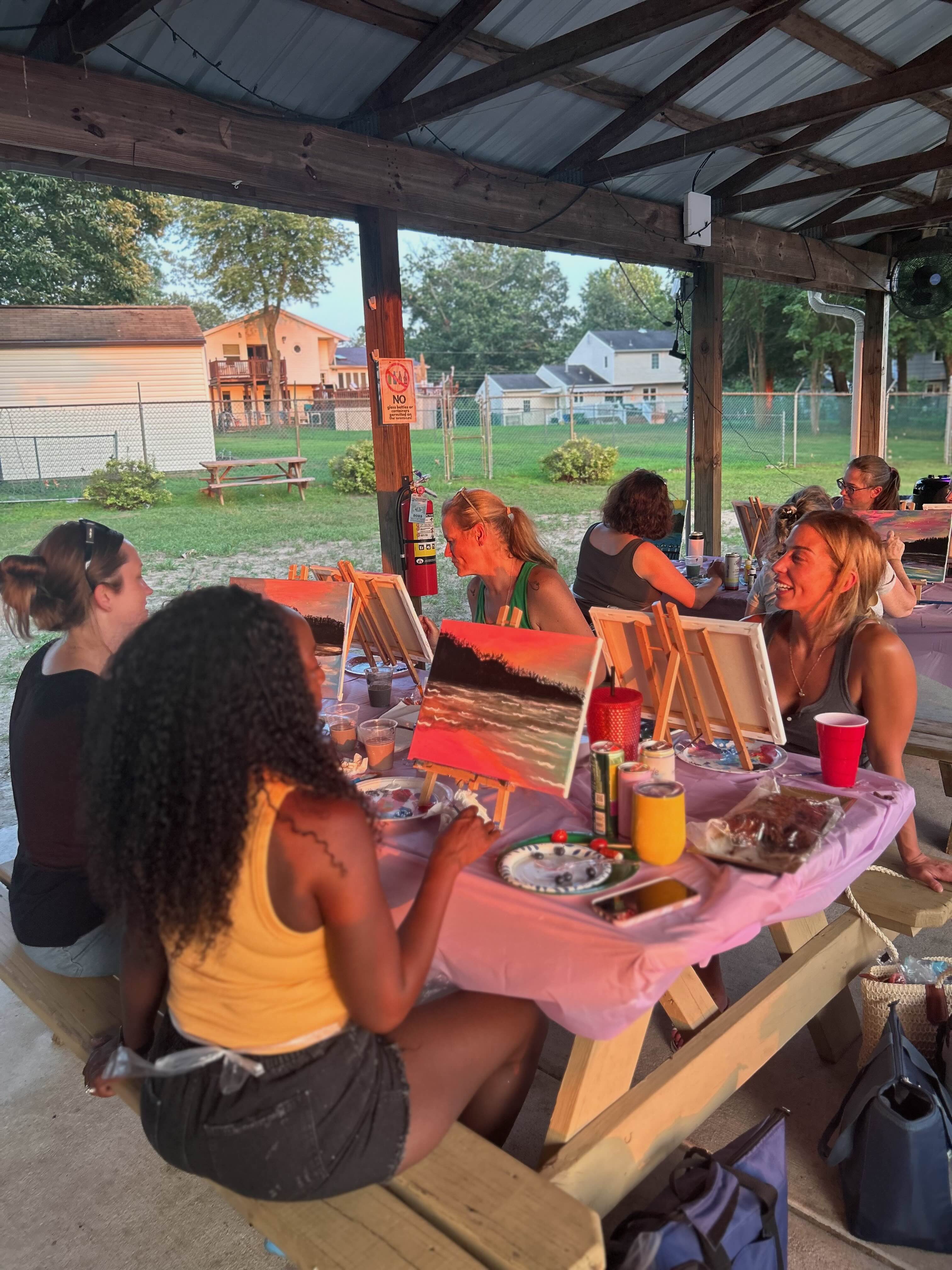 Group of women painting on canvases at an outdoor pavilion during sunset.