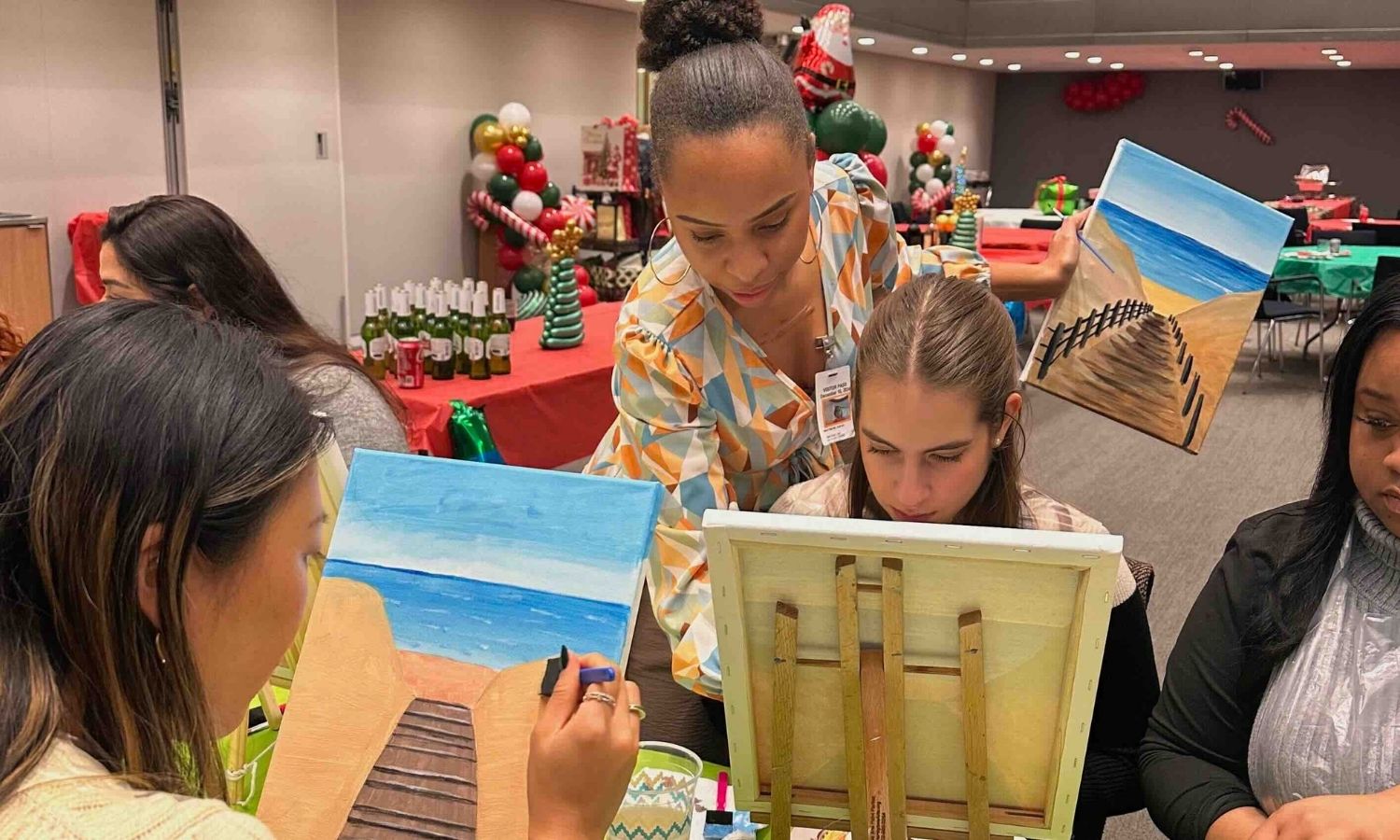 Women participating in a painting class with beach path paintings and holiday decorations in the background.