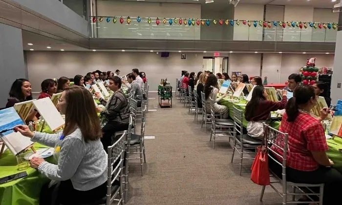Group of people seated at tables in a room, painting on canvases during an art class or paint party.