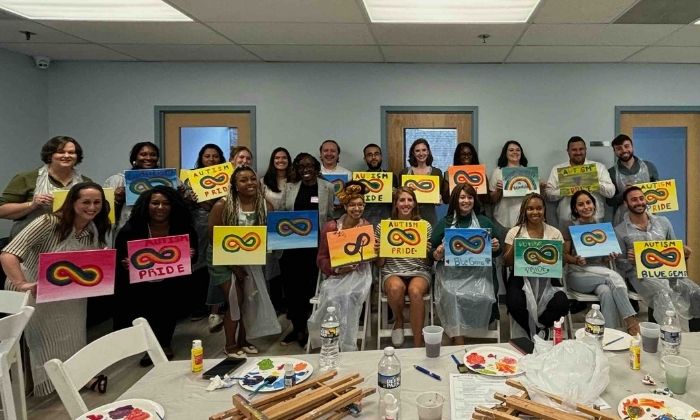 Group of people in a room holding colorful paintings of the autism pride infinity symbol.