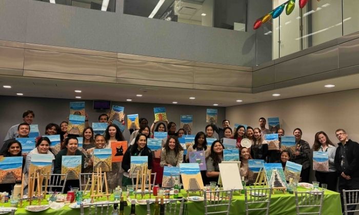 Group of diverse people holding up paintings of a beach boardwalk indoors after a painting party.