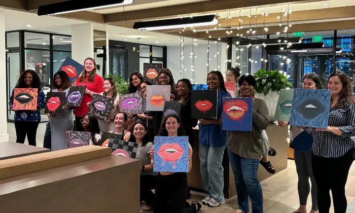 Group of diverse women indoors holding paintings of colorful lips on canvases at an art event.