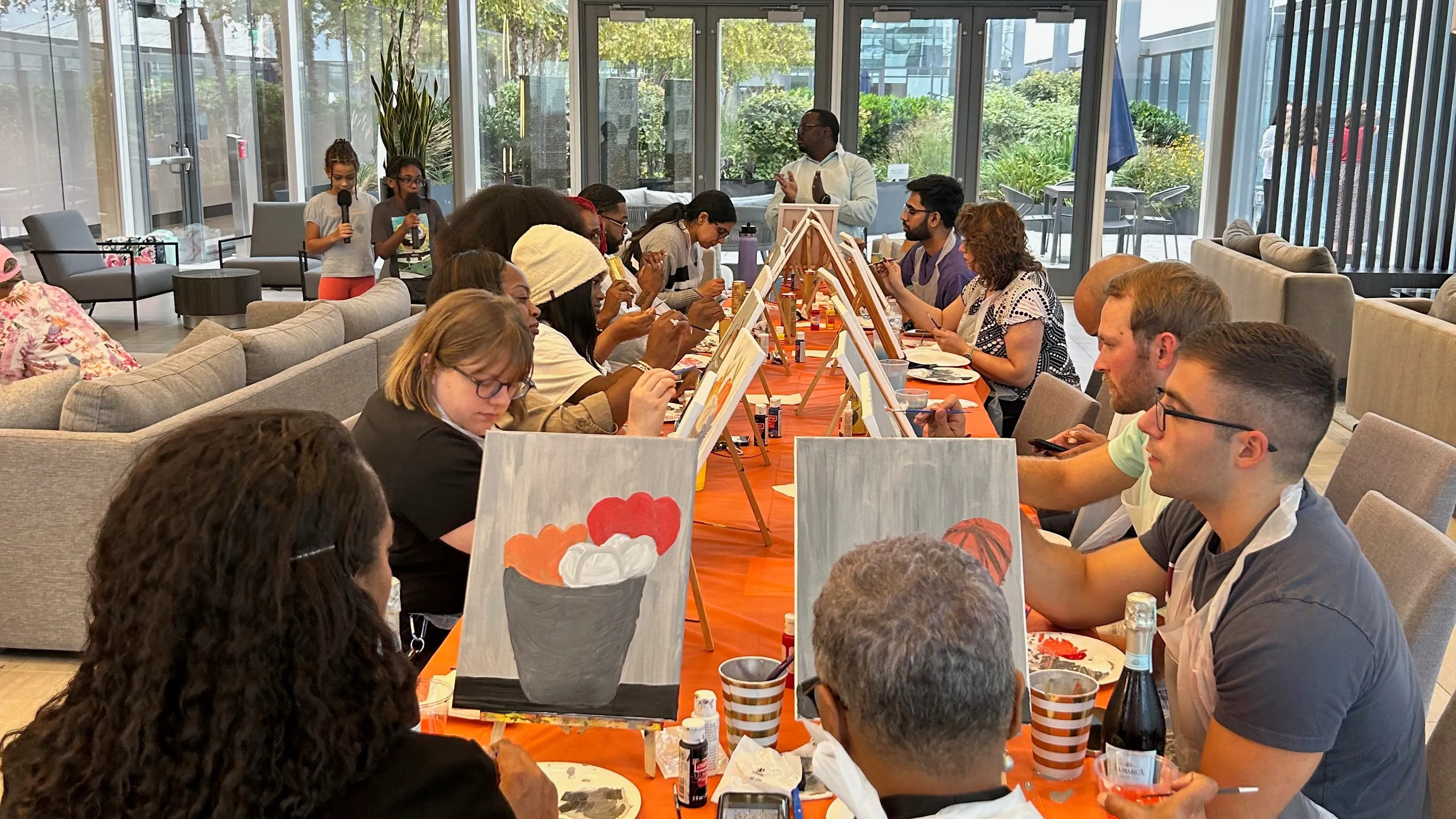 Group of adults seated at a long table painting on canvases during a social art class indoors.