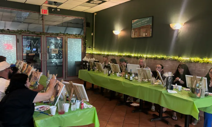 Group of people seated at two tables with green tablecloths painting on canvases in a cozy room.