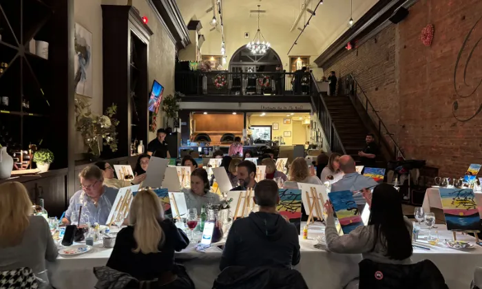 Group of people sitting at long table painting on easels in a cozy art studio with brick walls.
