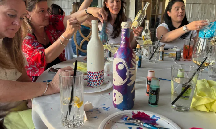 Group of women painting decorative bottles at a table with art supplies and drinks.