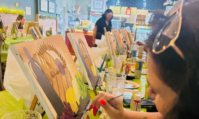 People painting on canvases at a sip and paint event in a brightly lit room with art supplies on tables.