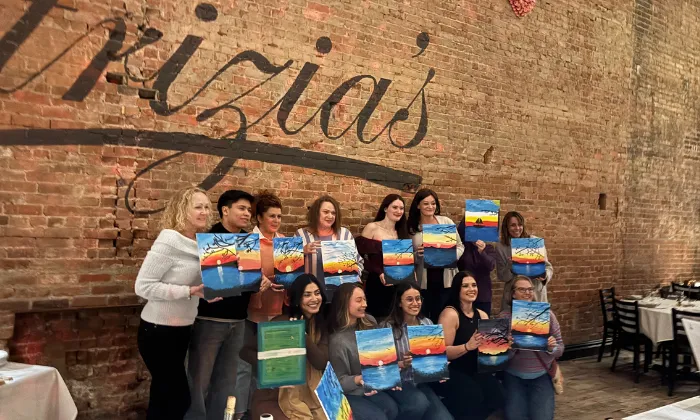 Group of people posing with colorful sunset paintings inside a restaurant with a brick wall reading 'pizia's'.
