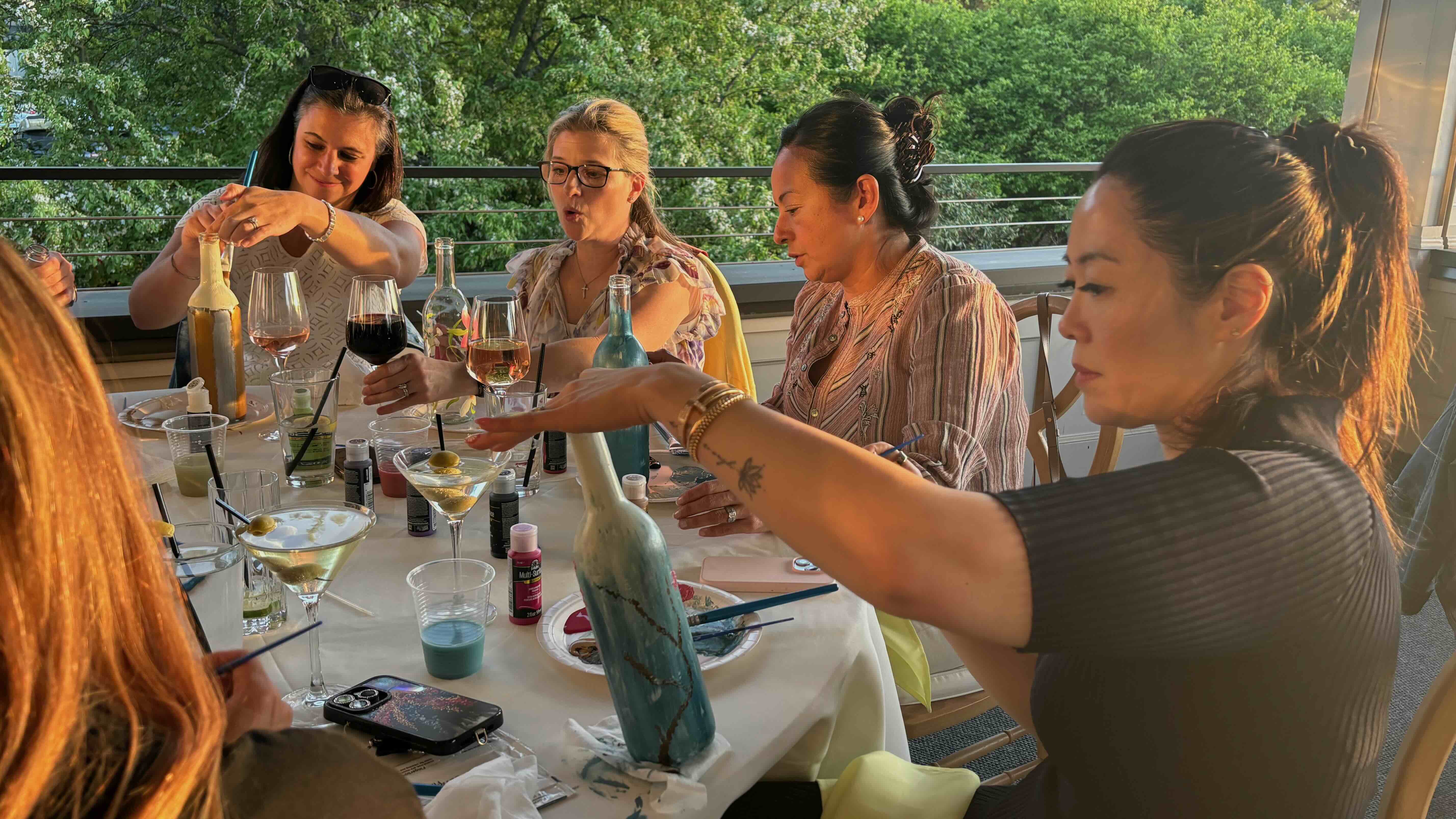 Women painting bottles together at a table with drinks and art supplies on an outdoor patio.