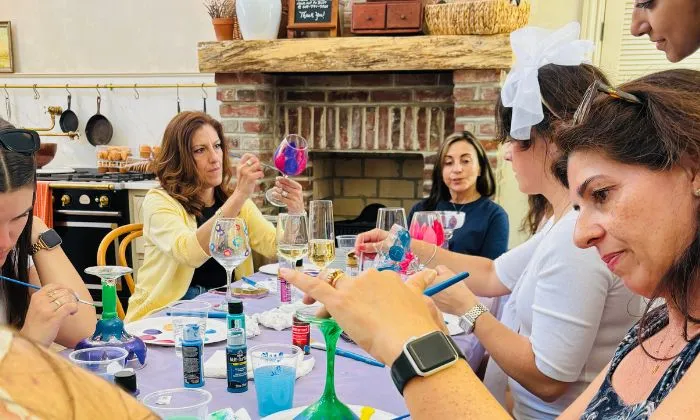 Group of nine women standing in a wine tasting room holding paintings of lighthouses by the sea.