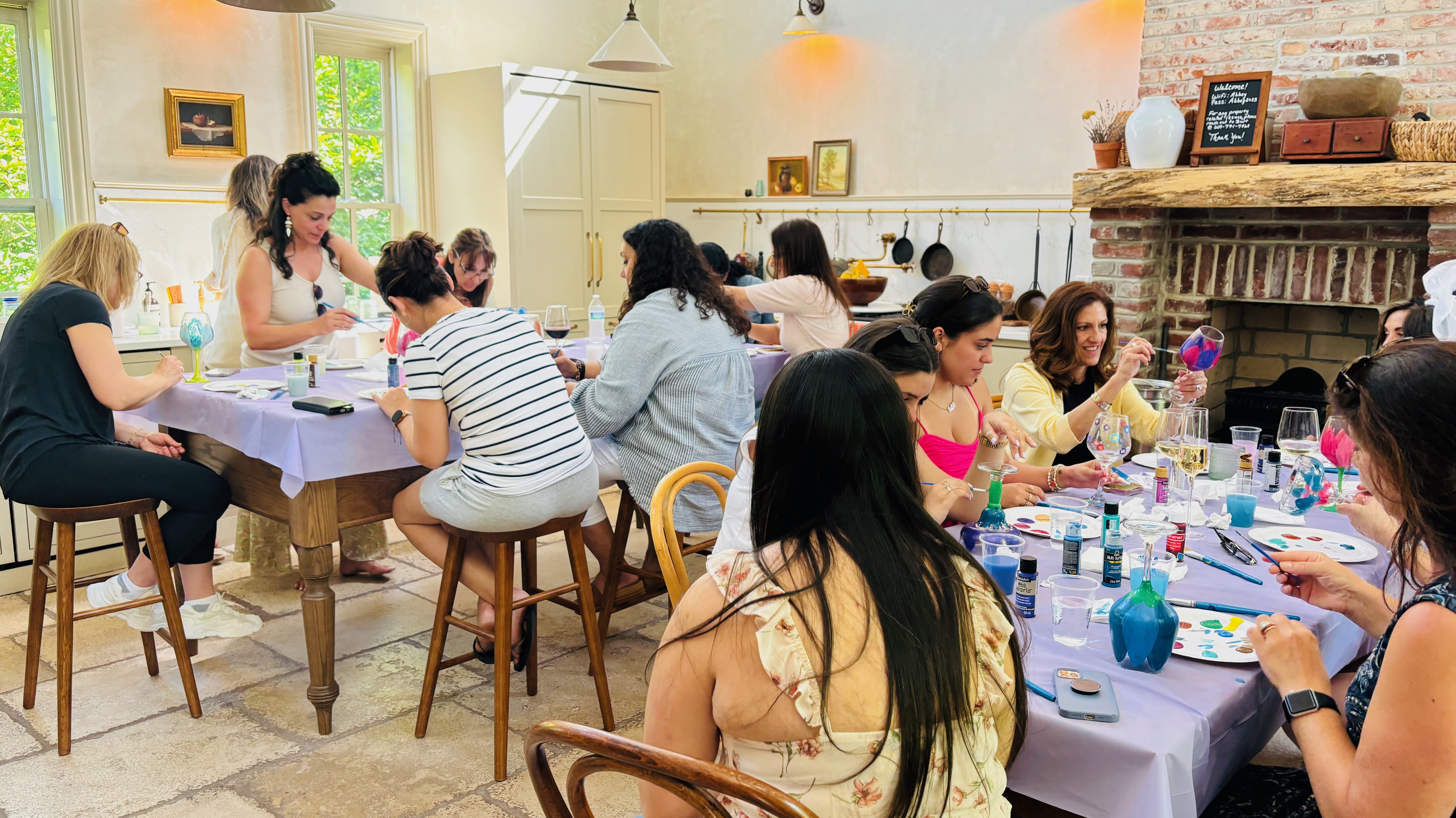 Women painting bottles together at a table with drinks and art supplies on an outdoor patio.