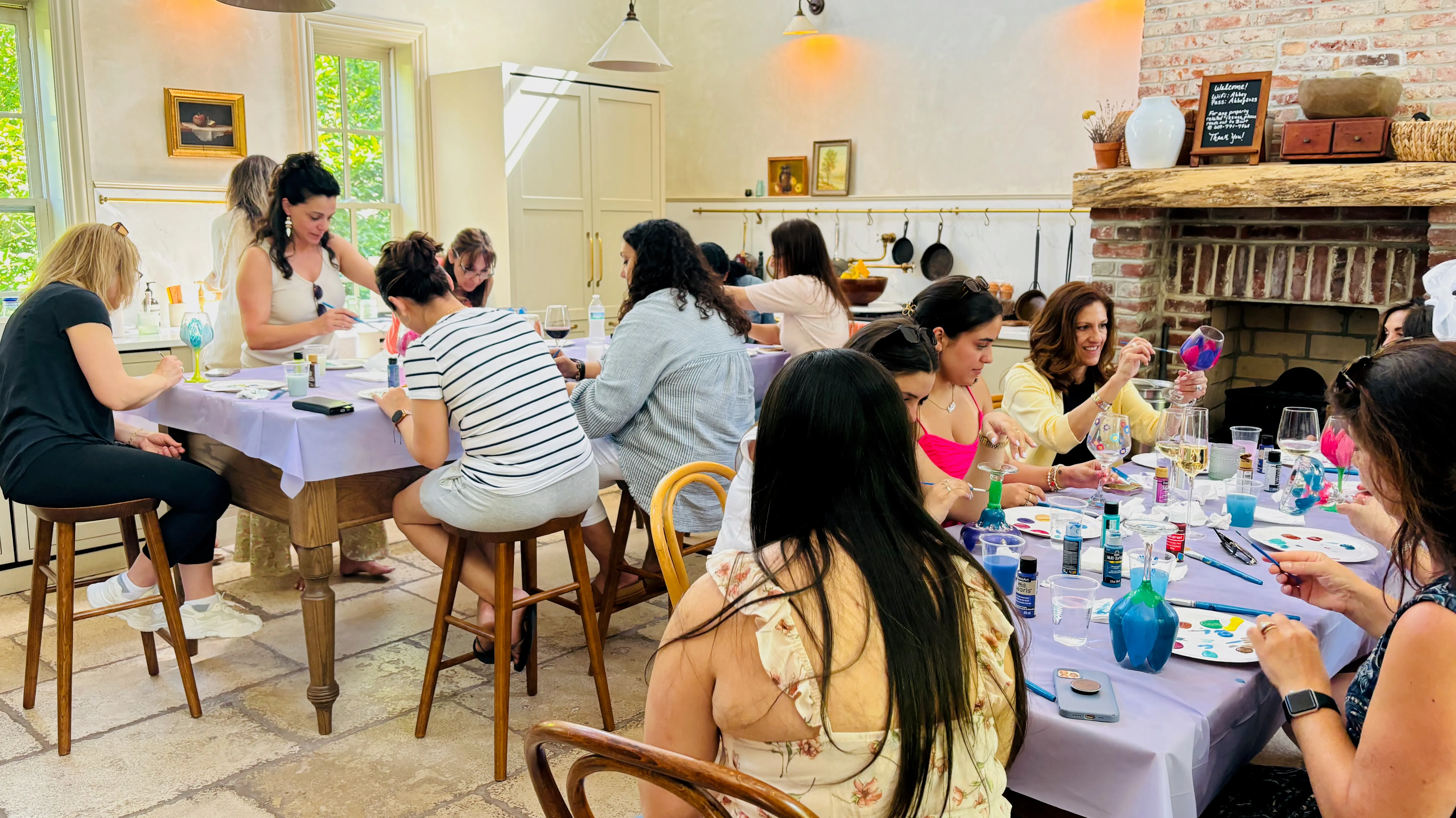 Women painting bottles together at a table with drinks and art supplies on an outdoor patio.