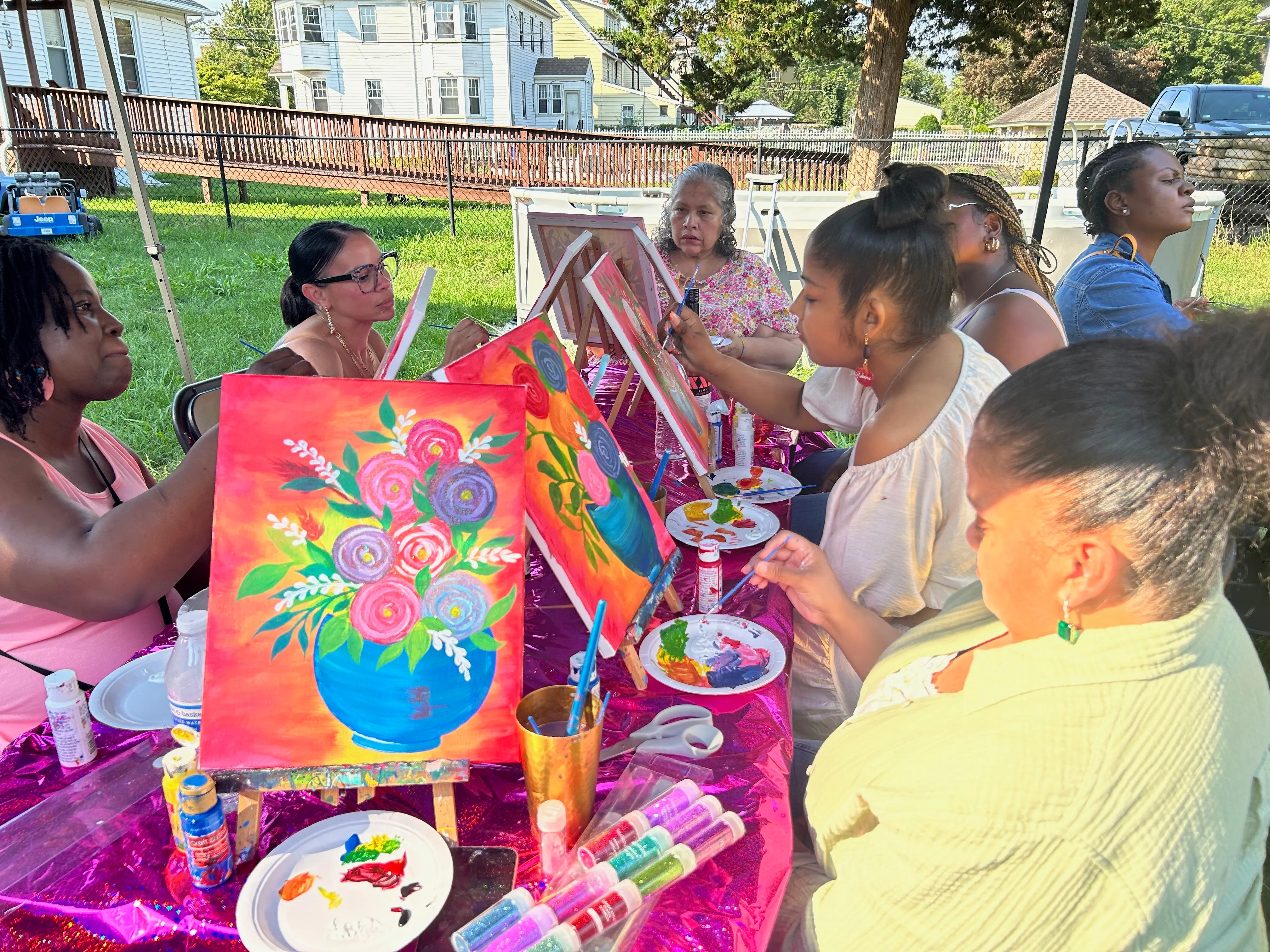 Group of women sitting outdoors painting colorful floral artworks on canvases at a decorated table.