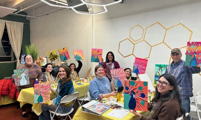 Group of people at a studio paint party showing their colorful peacock paintings and smiling.