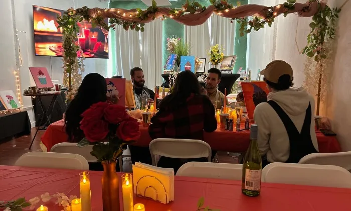 Group of people painting together at a warmly lit table decorated with flowers and candles in a paint and sip studio.