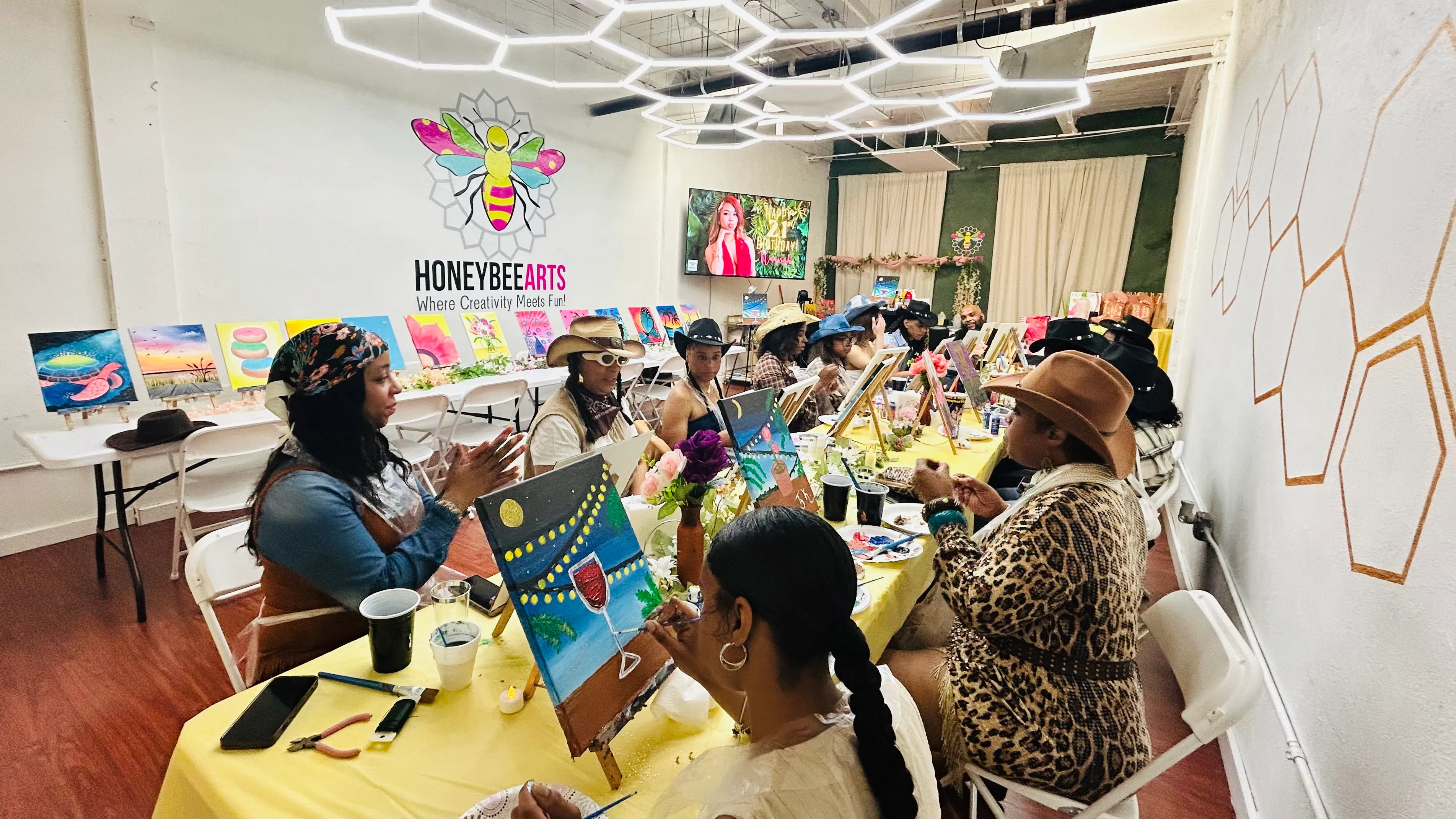 Group of people painting on canvases at HoneyBee Arts studio with yellow tablecloths and wall art.