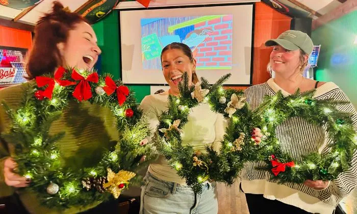 Three women smiling and holding decorated Christmas wreaths with lights and ornaments indoors.