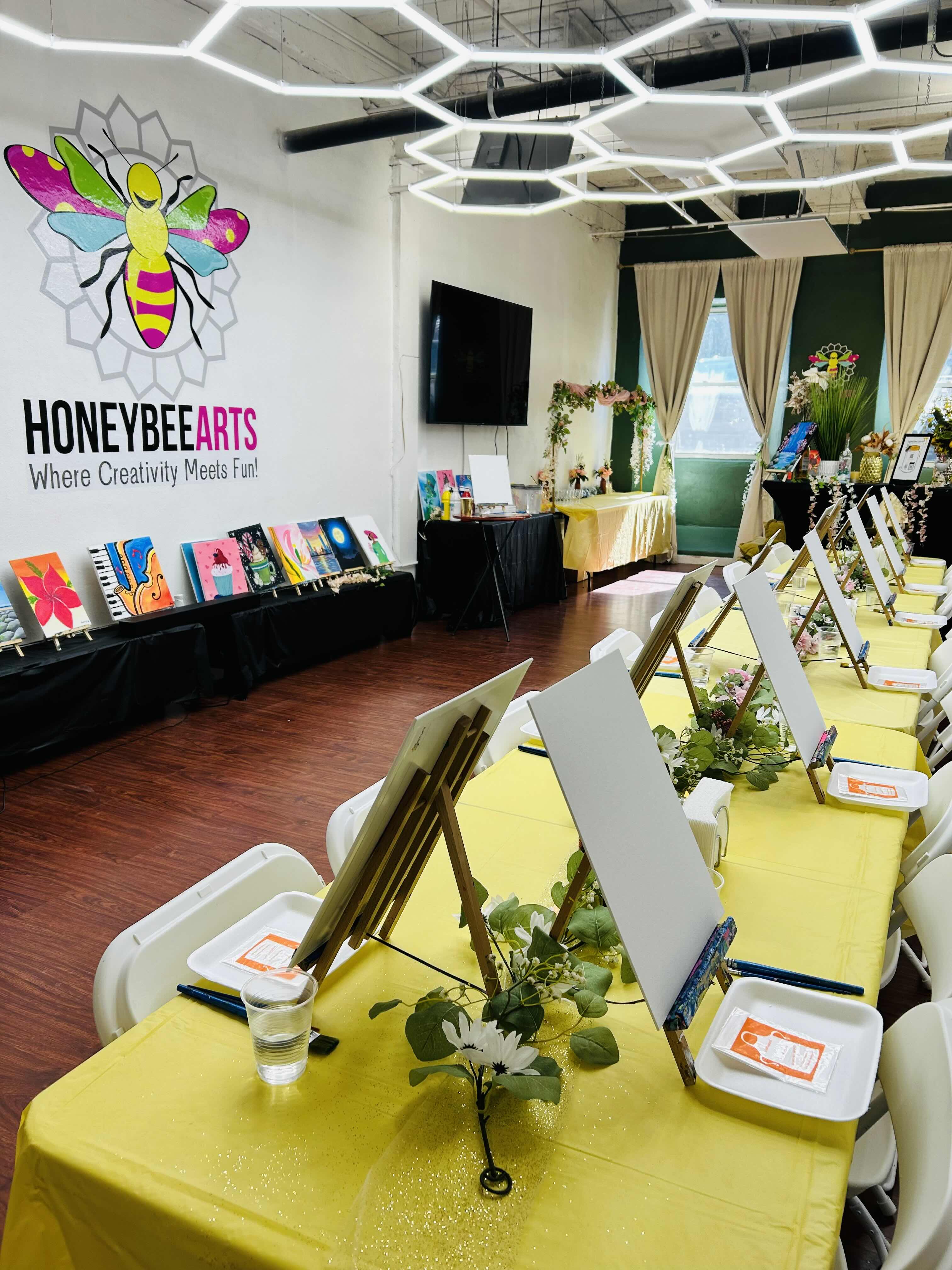 Bright art studio with yellow tables, blank canvases, paintings displayed, and a honeybee arts wall logo.