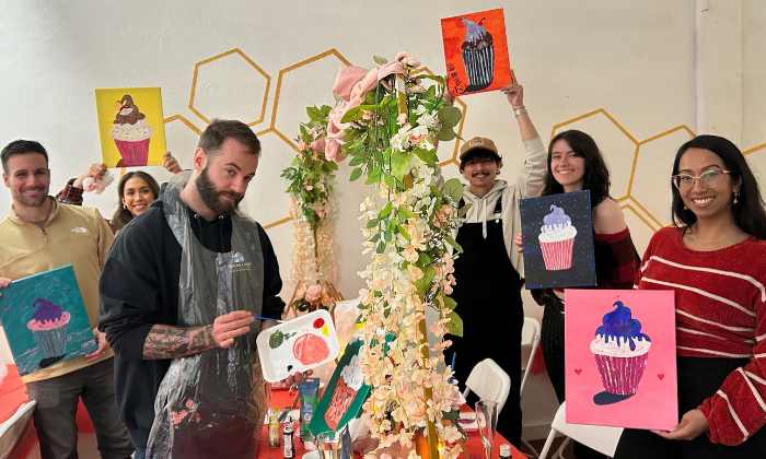 Group of diverse people holding painting canvases with blue waterfalls and cherry blossoms, posing in front of a colorful bee mural.